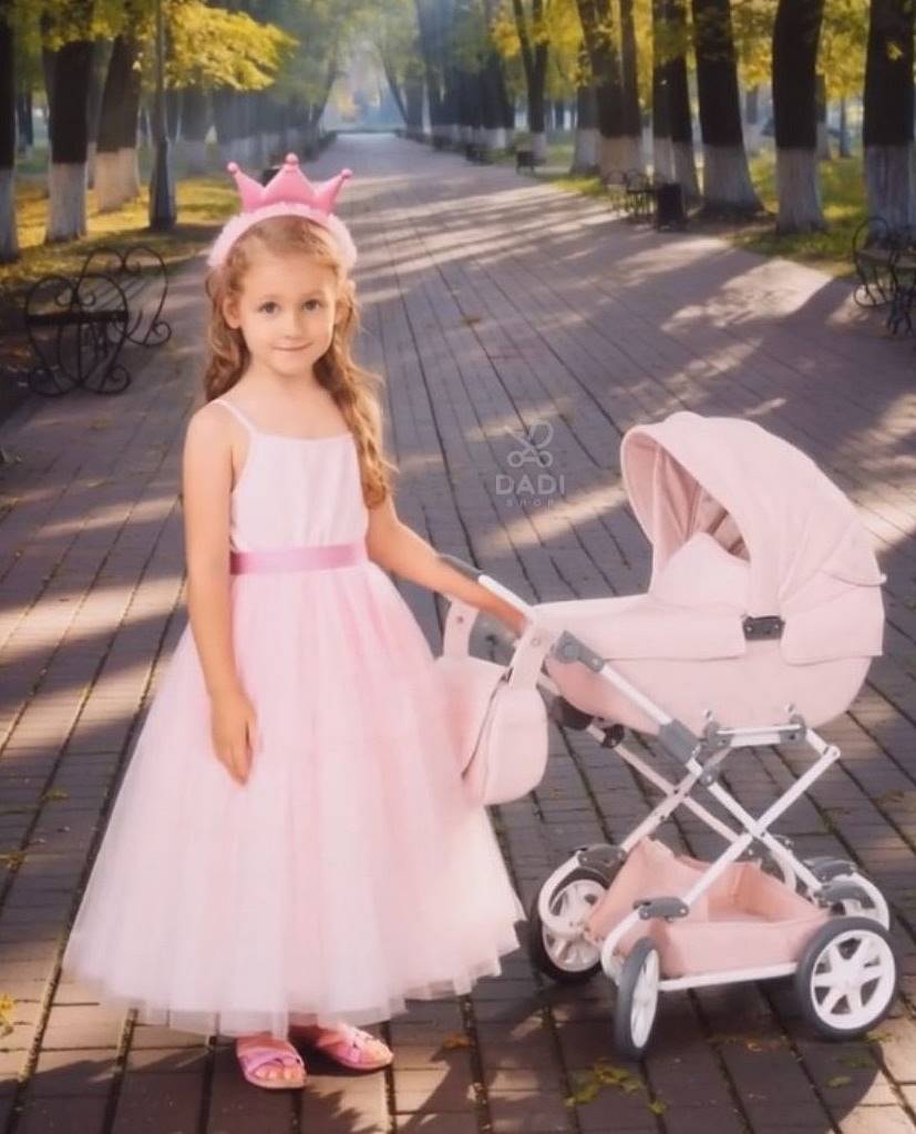 Dziewczynka w różowej sukience i koronie pozuje obok wózka dla lalek Tako Imperial Mini w kolorze pudrowego różu – idealny wybór dla małych księżniczek.